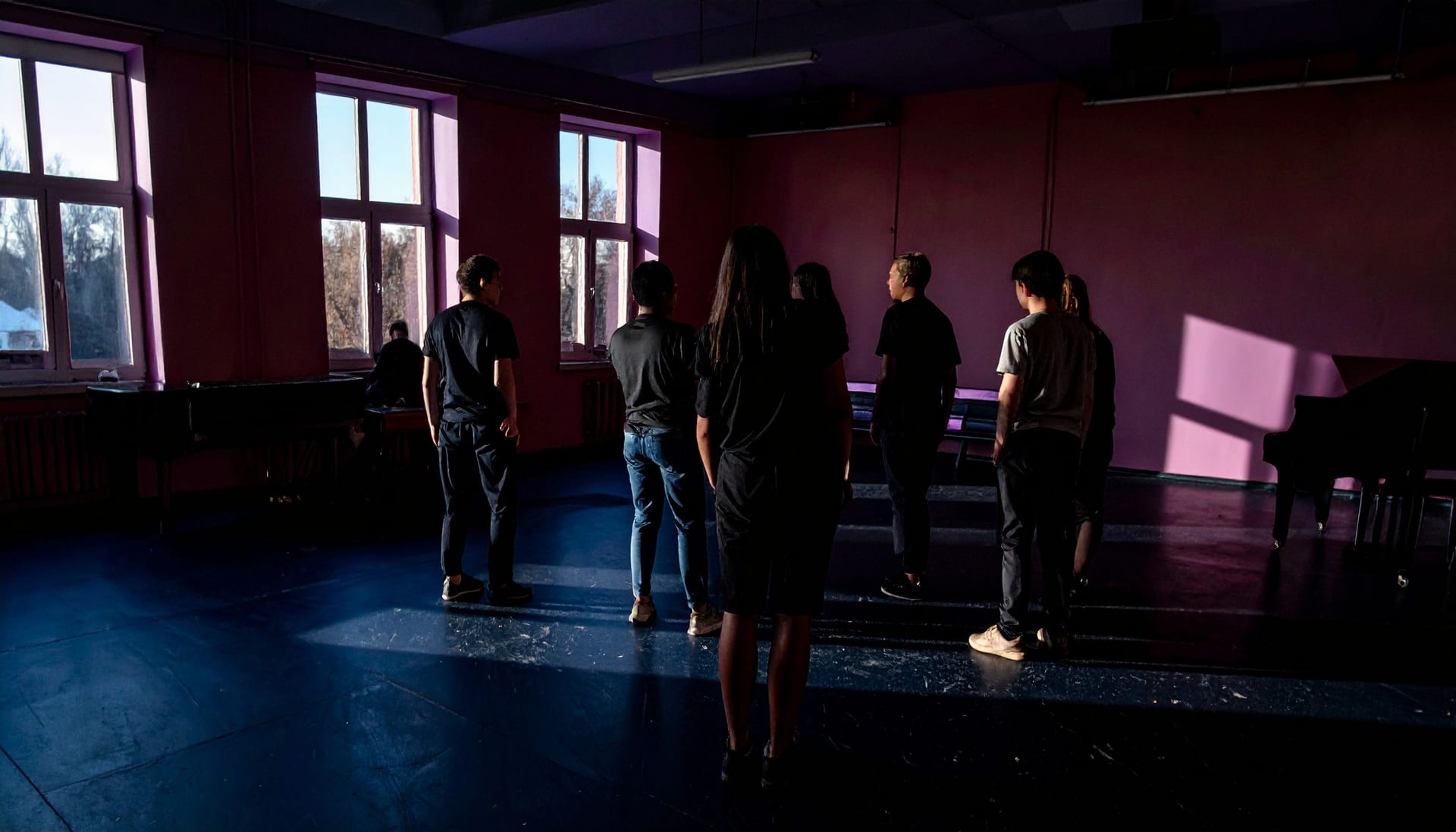 A high-school theatre rehearsal room in late afternoon, students standing in a loose circle silhouetted against tall windows.