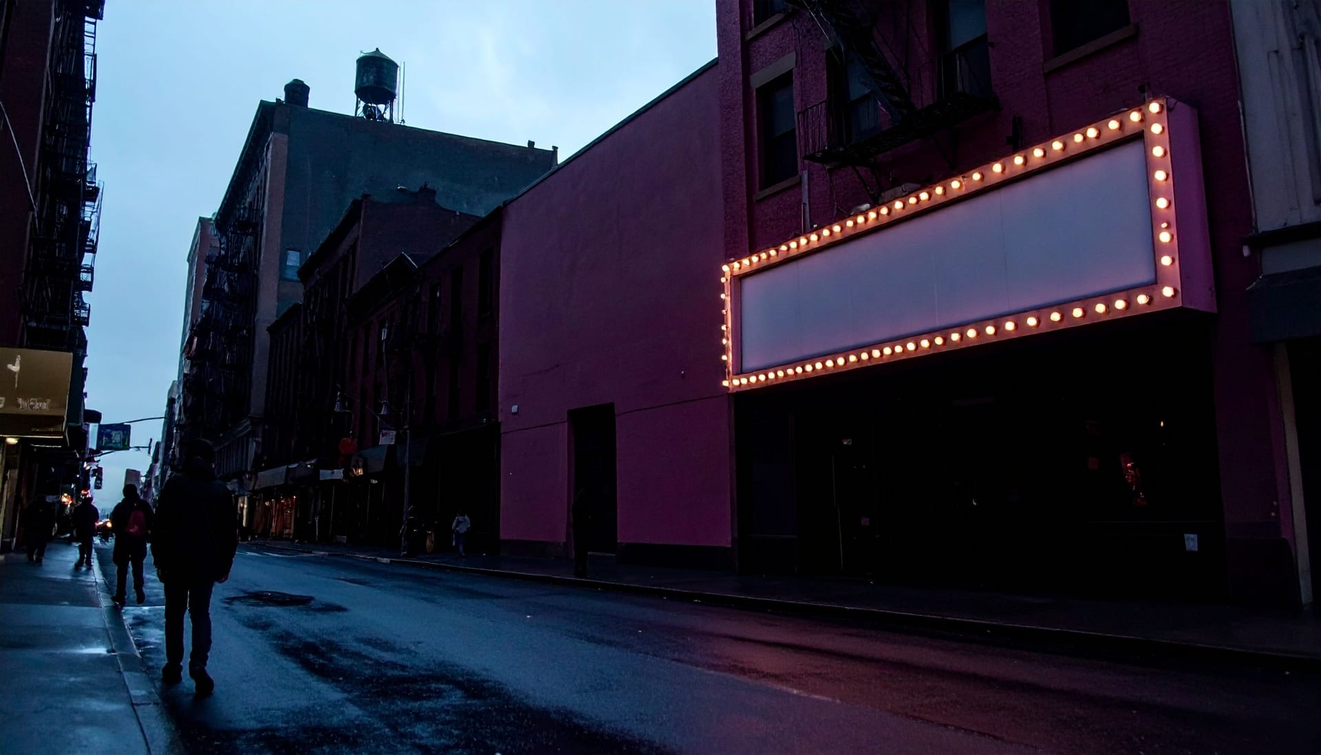 A New York theatre marquee at dusk seen from across the street, bulbs unlit around blank panels, water towers on the rooflines above.