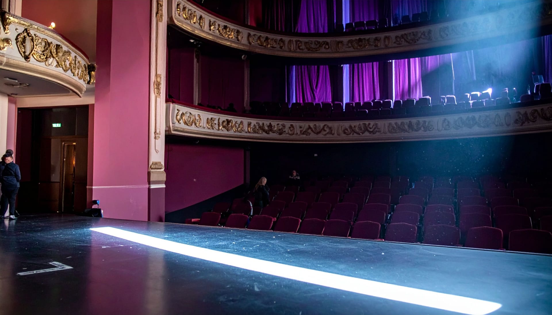 An empty theatre seen from the edge of the stage, rows of red seats receding into darkness under a single work light.