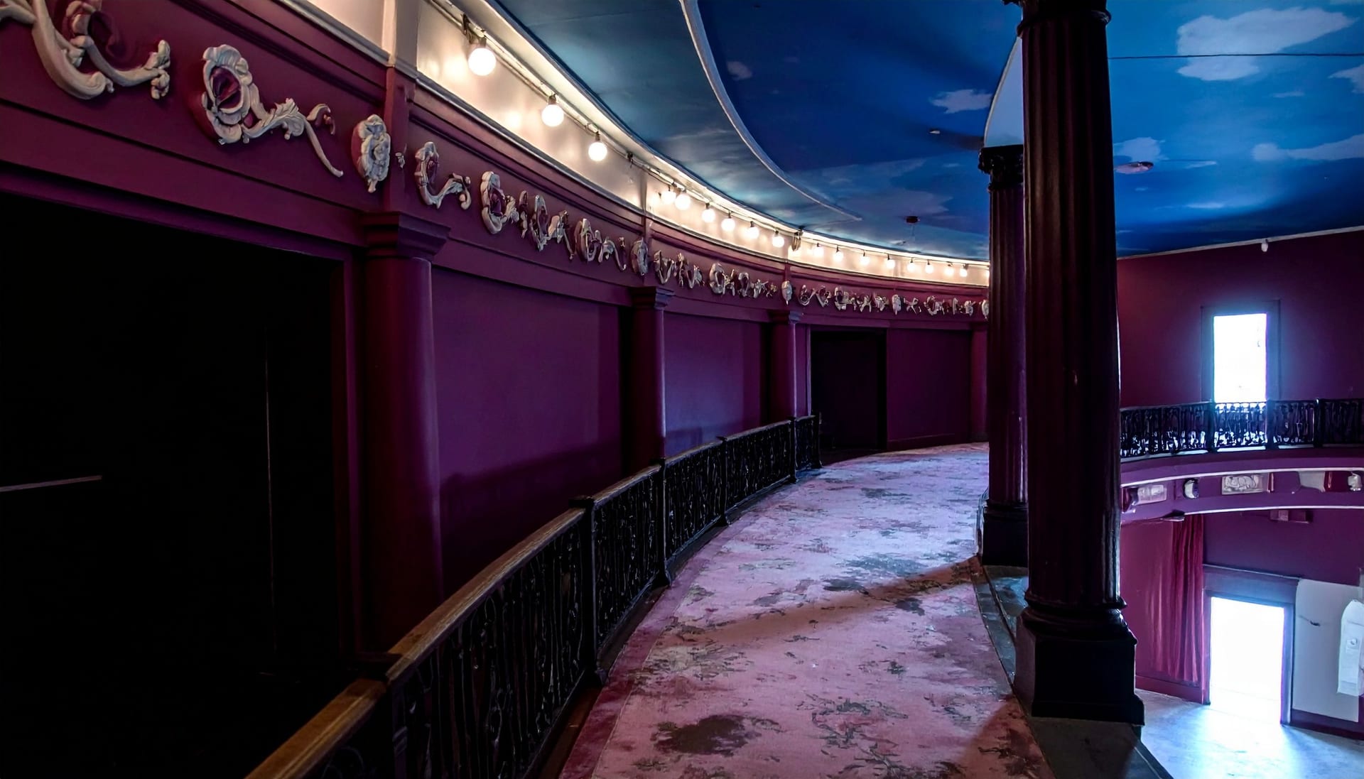 The curving mezzanine promenade of an old Broadway theatre in the evening, deep plum walls and painted plasterwork above a worn rose carpet, a row of small bulbs following the balcony rail and a painted cloud ceiling overhead.
